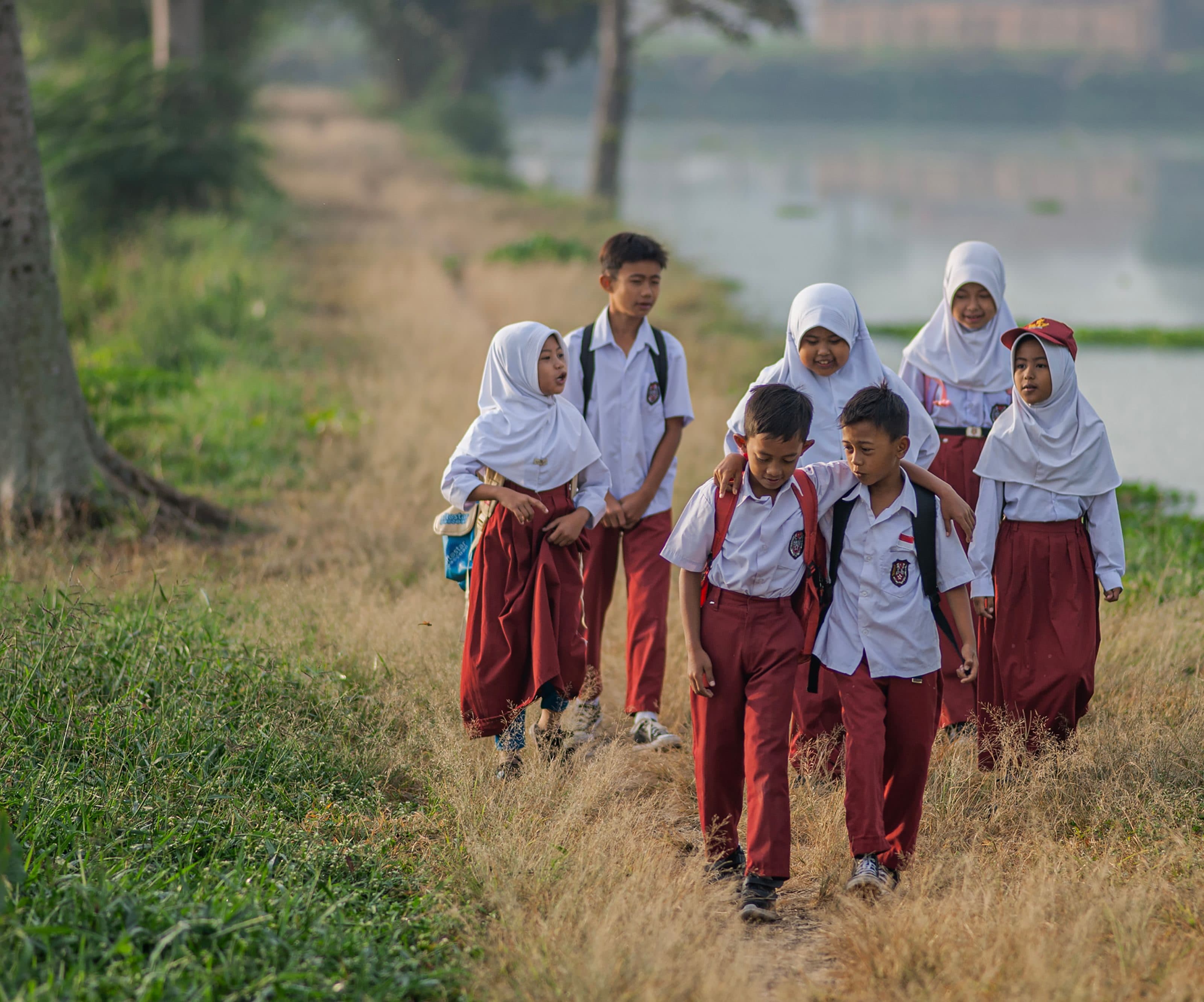 Tujuh anak sekolah dasar berseragam putih dan merah berjalan beriringan di jalan setapak tepi danau pada pagi hari